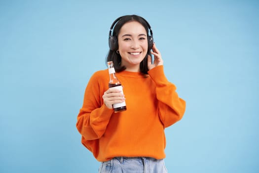 Beautiful asian woman listens music in headphones, drinks coke from bottle and smiles at camera, blue background.