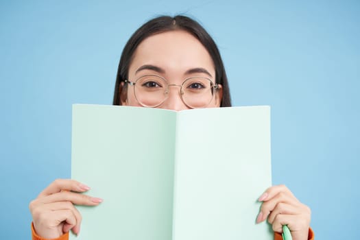 Portrait of cute japanese woman in glasses, peeking from notebook, covers face with her notes, smiles with eyes, blue background.