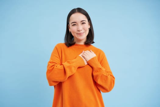 Care and love. Asian woman holds hands on heart and smiles, looks with tenderness and warmth at camera, stands over blue background.