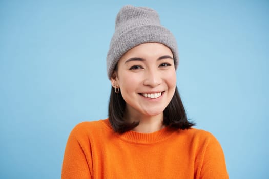 Close up portrait of smiling, beautiful young woman, asian girl in hat, looking happy and candid at camera, standing over blue background.