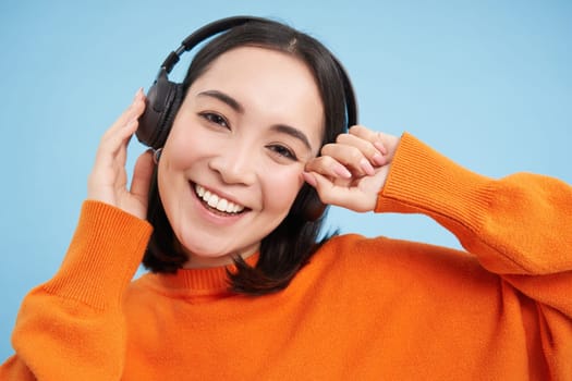 Close up of beautiful asian woman in headphones, smiles and listens music, enjoys podcast, stands over blue background.