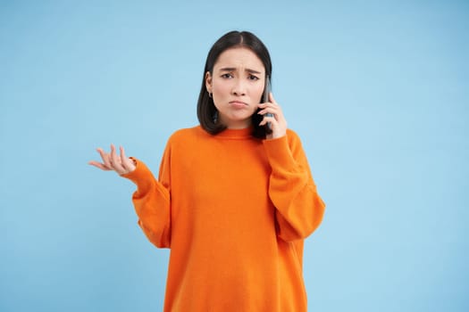 Sad and confused asia girl shrugs, talks on cellphone, answers telephone call with puzzled face, stands over blue background.