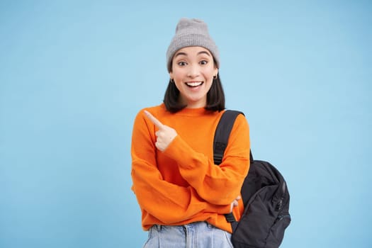 Enthusiastic asian woman points left, shows promo offer, holds backpack, stands over blue background.