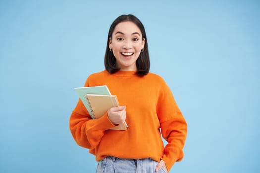 Young asian student with notebooks, standing in orange sweatshirt, looking at camera, posing over blue background.