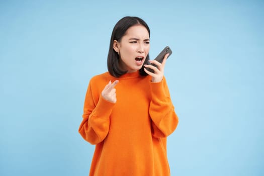 Angry asian woman yells at mobile phone, shouts in smartphone with furious face, stands over blue background.