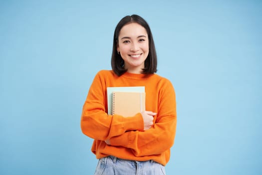 Education and students. Happy asian woman, holding notebooks and laughing, smiling at camera, enjoys going to University or College, blue background.