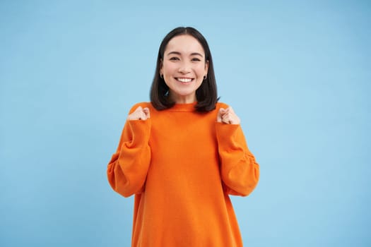 Cheerful asian girl champion, rooting for smth, raising hands up joyful, smiling and celebrating, winning, chanting with upbeat face, blue background.