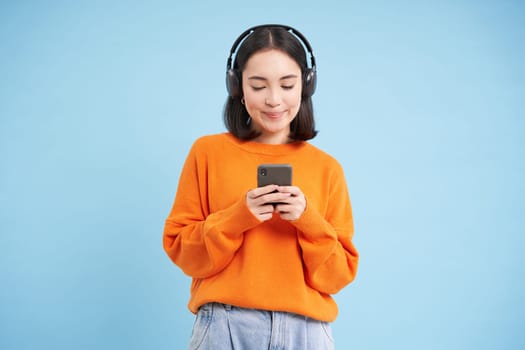 Happy young modern woman with headphones and smartphone, listening to music on mobile phone, enjoys favourite podcast, blue background.