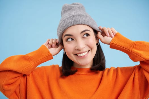 Close up portrait of smiling asian woman in warm hat, looking happy and cute at camera, has clear natural skin, stands over blue background.