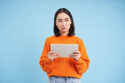 Woman with digital tablet thinking, looking aside with thoughtful face expression, standing over blue background.