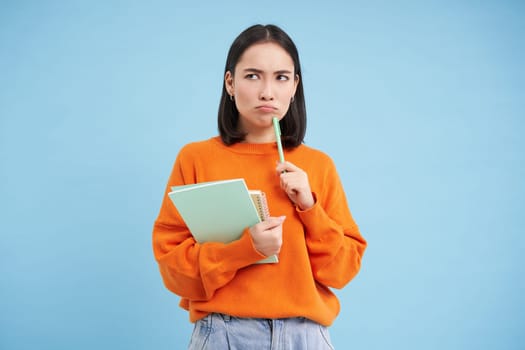 Asian girl, student with thinking face, pondering smth with serious face, holding pencil and notebooks, standing over blue background.