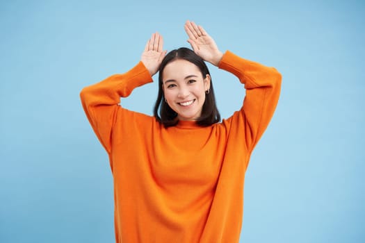 Funny young woman shows ears gesture above head and dancing, looking cute and silly, playing, standing over blue background.