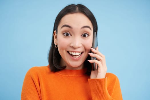 Cheerful asian girl talks on mobile phone, speaks with someone on telephone, stands over blue background. Technology concept