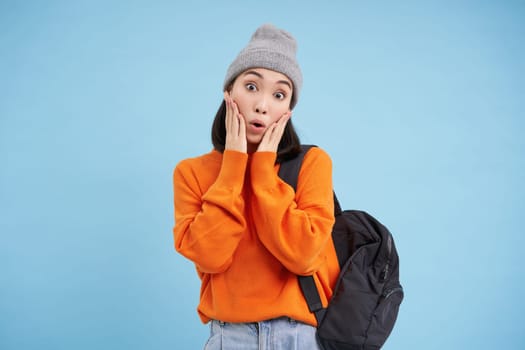 Stylish young asian woman in warm hat, walking with backpack, going somewhere with bag, standing over blue studio background.