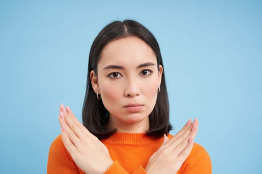 Close up portrait of serious asian woman, shows stops sign, disapproves something, refuses or rejects, stands over blue background.