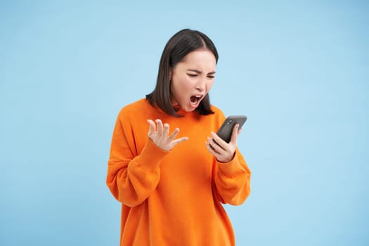 Angry korean girl swearing and shouting at mobile phone, looks outraged, furious while reading news on smartphone, standing over blue background.