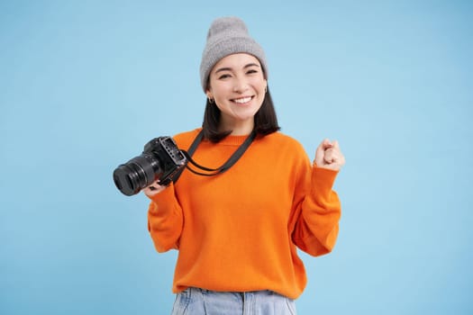 Stylish asian girl with digital camera, taking pictures. Woman photographer smiling, standing over blue background.