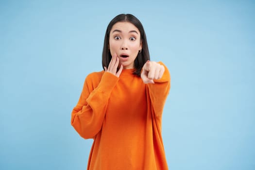 Looks at that, omg. Shocked asian woman points finger at camera with dropped jaw, sees something unbelievable, impressed by you, stands over blue background.