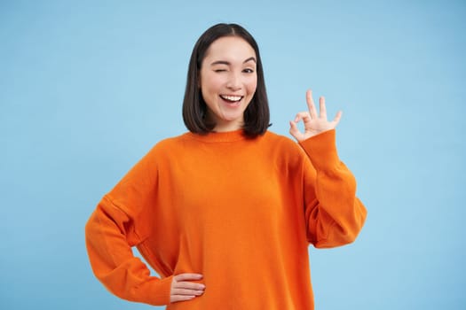 Alright, very good. Smiling winking girl, asian woman shows okay sign in approval, satisfied with smth, stands over blue background.