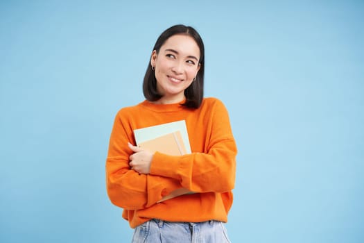 Education and students. Happy asian woman, holding notebooks and laughing, smiling at camera, enjoys going to University or College, blue background.