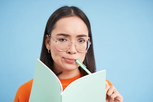 Portrait of asian girl, student in glasses, thinking, writing homework, making notes, holding pen, frowns and looks puzzled, blue background.