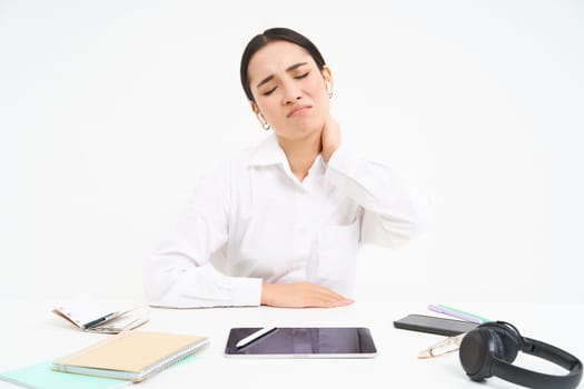 Tired asian woman, office lady sits behind her desk, has back pain, massaging her shoulder, tired after work, white background.