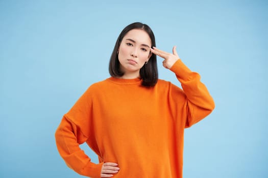 Portrait of asian woman annoyed by something, holds two fingers near head temple, bang bang gesture, stands over blue background.