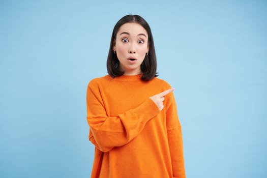 Surprised korean woman, pointing right, showing promo banner with amazed face expression, stands over blue background. Copy space