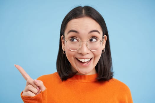 Portrait of japanese woman in glasses, points left, looks aside at banner, stands over blue studio background.