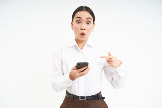 Business concept. Impressed asian corporate woman, pointing at mobile phone with surprised face, amazed by smartphone app, standing over white background.