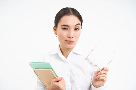 Confident young woman, office worker with glasses, holds notebooks, looks thoughtful, thinking, standing isolated on white background.