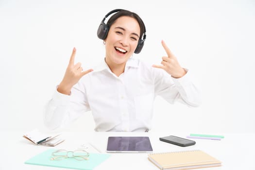 Happy and excited korean woman in headphones, manager sits at desk in office with digital tablet and work documents, shows rock n roll sign, listens music.
