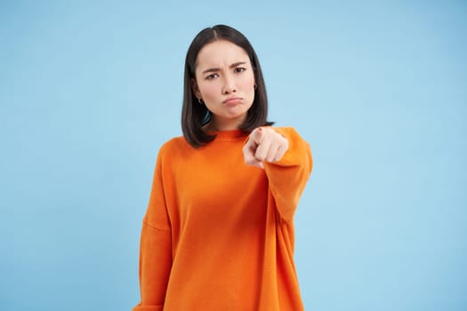 Angry asian woman pointing at camera with blame, blaming you, accusation gesture, standing over blue background.