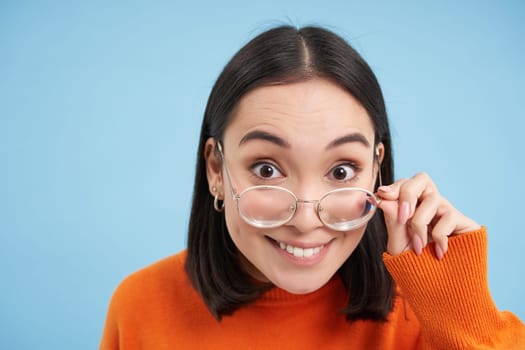 Portrait of brunette japanese woman in glasses, smiling and looking with happy face at camera, tries prescribed eyewear, blue background.