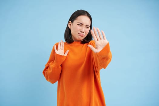 Stop it, no photos please. Upset asian girl blocks camera with hands, doesnt want to take pictures, stands over blue background.