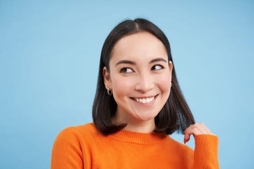 Close up portrait of asian girl with perfect healthy smile and natural beautiful face, looks happy at camera, stands over blue background.