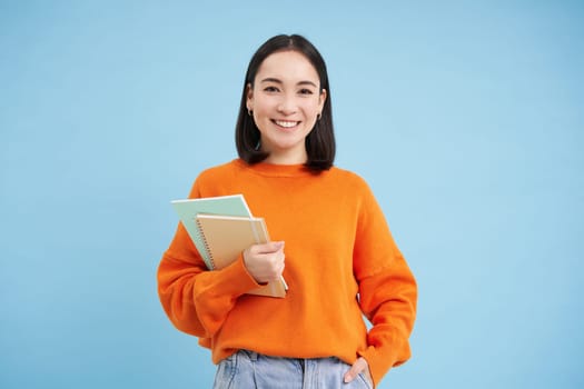 Confident smiling korean woman with notebooks, University student posing against blue background.