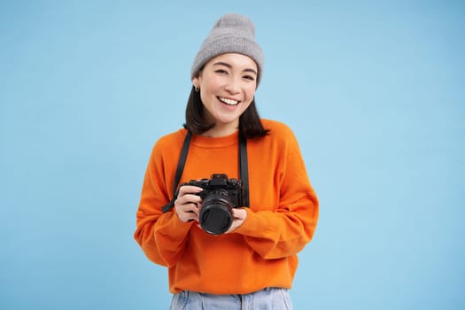 Stylish asian girl with digital camera, taking pictures. Woman photographer smiling, standing over blue background.