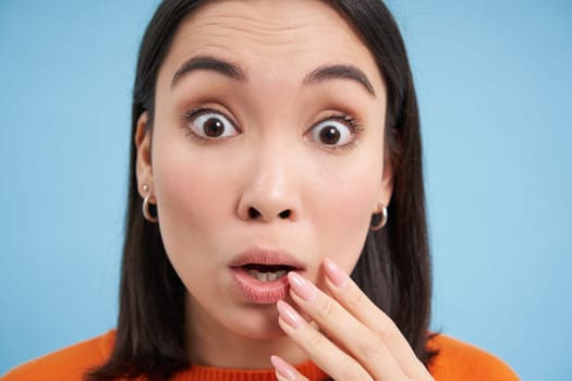 Close up portrait of amazed asian girl, looks with interest, listens to amazing story, impressed by something, stands over blue background.