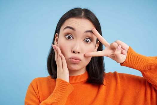 Close up of cute and silly japanese woman, shows peace sign and pouts, stands over blue background.