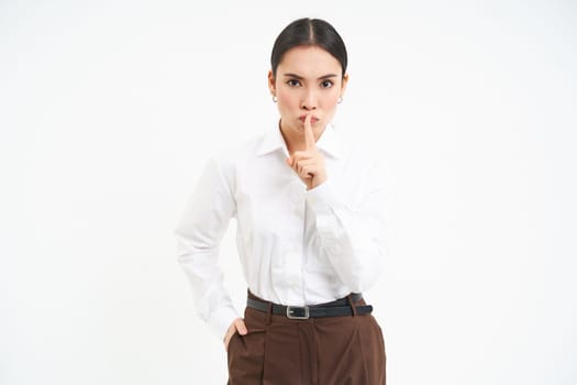 Strict and serious businesswoman shushing, puts finger on lips, tells to be quiet, hush gesture, stands over white background.