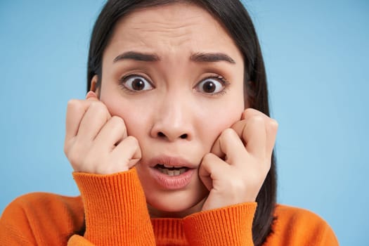 Portrait of scared, anxious young asian woman, looks horrified, stands over blue studio background.