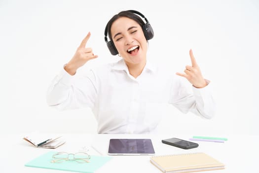 Happy and excited korean woman in headphones, manager sits at desk in office with digital tablet and work documents, shows rock n roll sign, listens music.