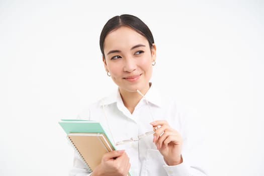Confident young woman, office worker with glasses, holds notebooks, looks thoughtful, thinking, standing isolated on white background.