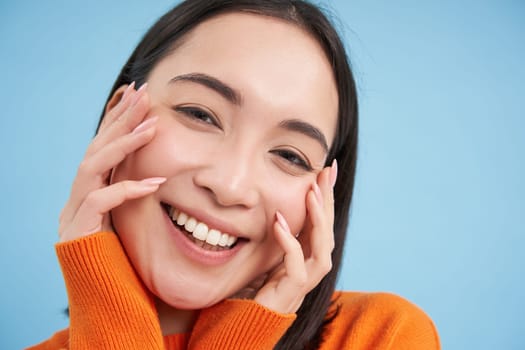 Close up portrait of beautiful japanese brunette girl, smiling and looking lovely at camera, gazing with care and tenderness, blue background.