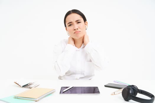 Tired asian woman, office lady sits behind her desk, has back pain, massaging her shoulder, tired after work, white background.