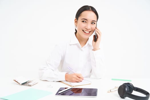 Business and company concept. Young asian woman, manager working in office, writing down information and talking with client on mobile phone, white background.