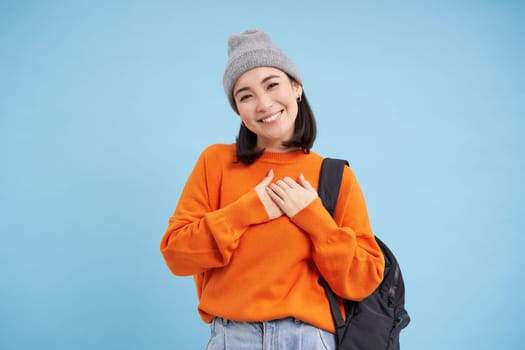 Lovely asian woman in warm hat, wears backpack, holds hands on heart and smiles, stands over blue background. Copy space