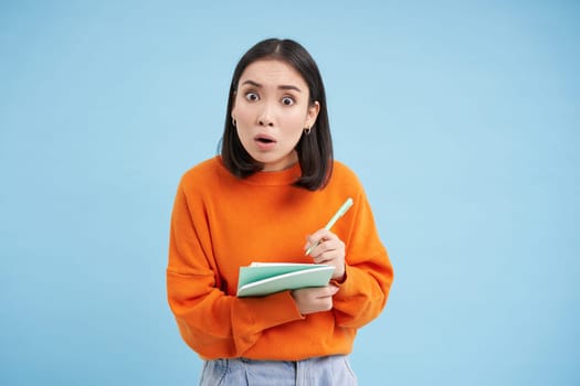 Education and students. Happy asian woman, holding notebooks and laughing, smiling at camera, enjoys going to University or College, blue background.
