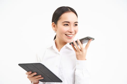 Portrait of confident korean businesswoman, holding digital tablet, sending voice message on smartphone, working on project, white background.
